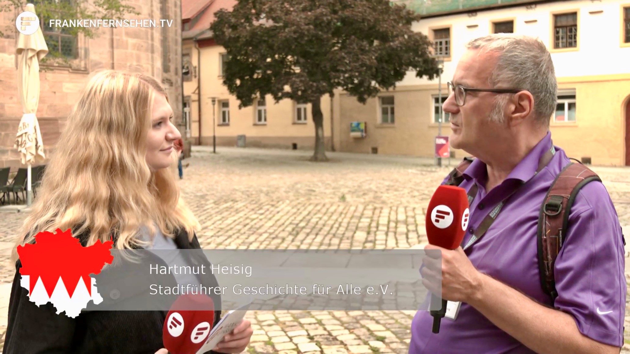 Am linken Bildrand steht eine Reporterin, ihr gegenüber beantwortet Rundgangsleiter Hartmut Heisig ihre Fragen und spricht in ein Mikrofon, im Hintergrund ein Stadtplatz mit Baum
