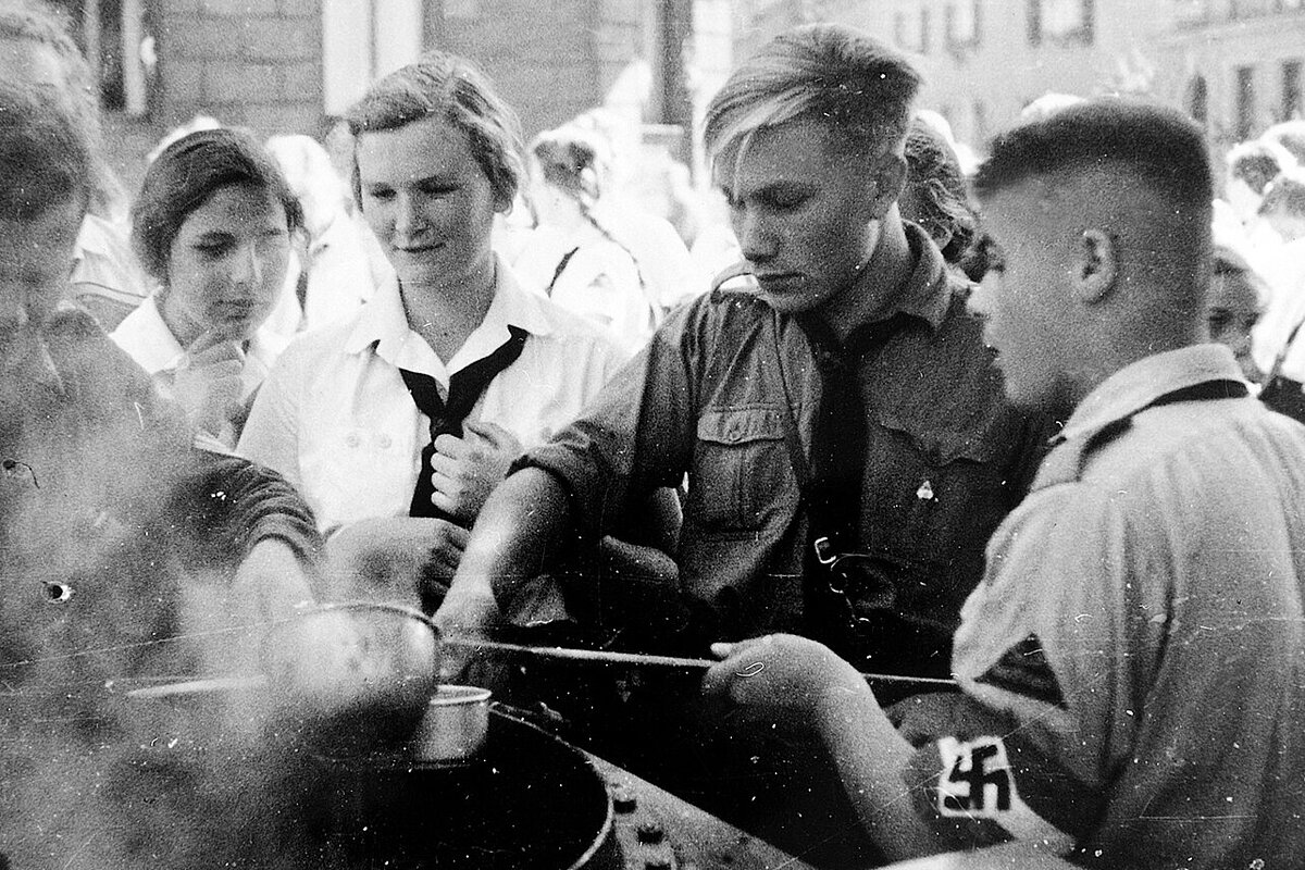 Schwarzweißaufnahme von jungen Männern und Frauen in nationalsozialistischen Uniformen, die um einen Topf Suppe stehen, die mit einer Kelle verteilt wird