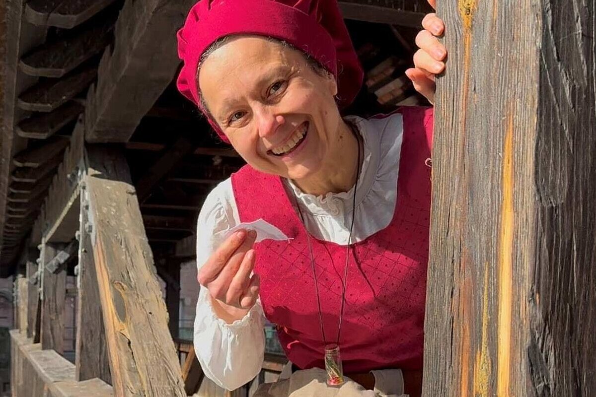 Frau in historischem Kostüm lächelt in die Kamera, im Hintergrund ein überdachter Holzsteg