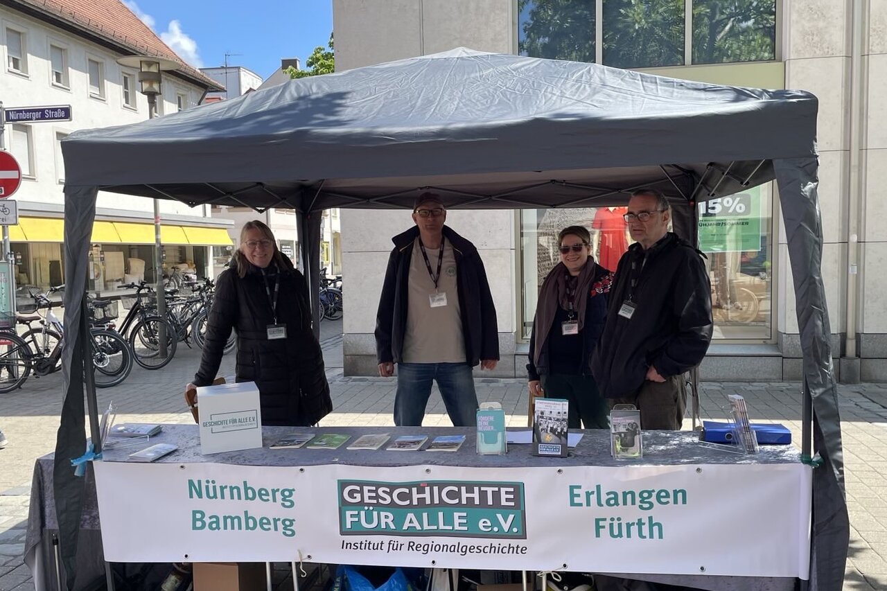 Zwei Frauen und zwei Männer in dunkler Kleidung stehen hinter einem Tisch mit Büchern und Infomaterial des Vereins unter einem grauen Pavillon