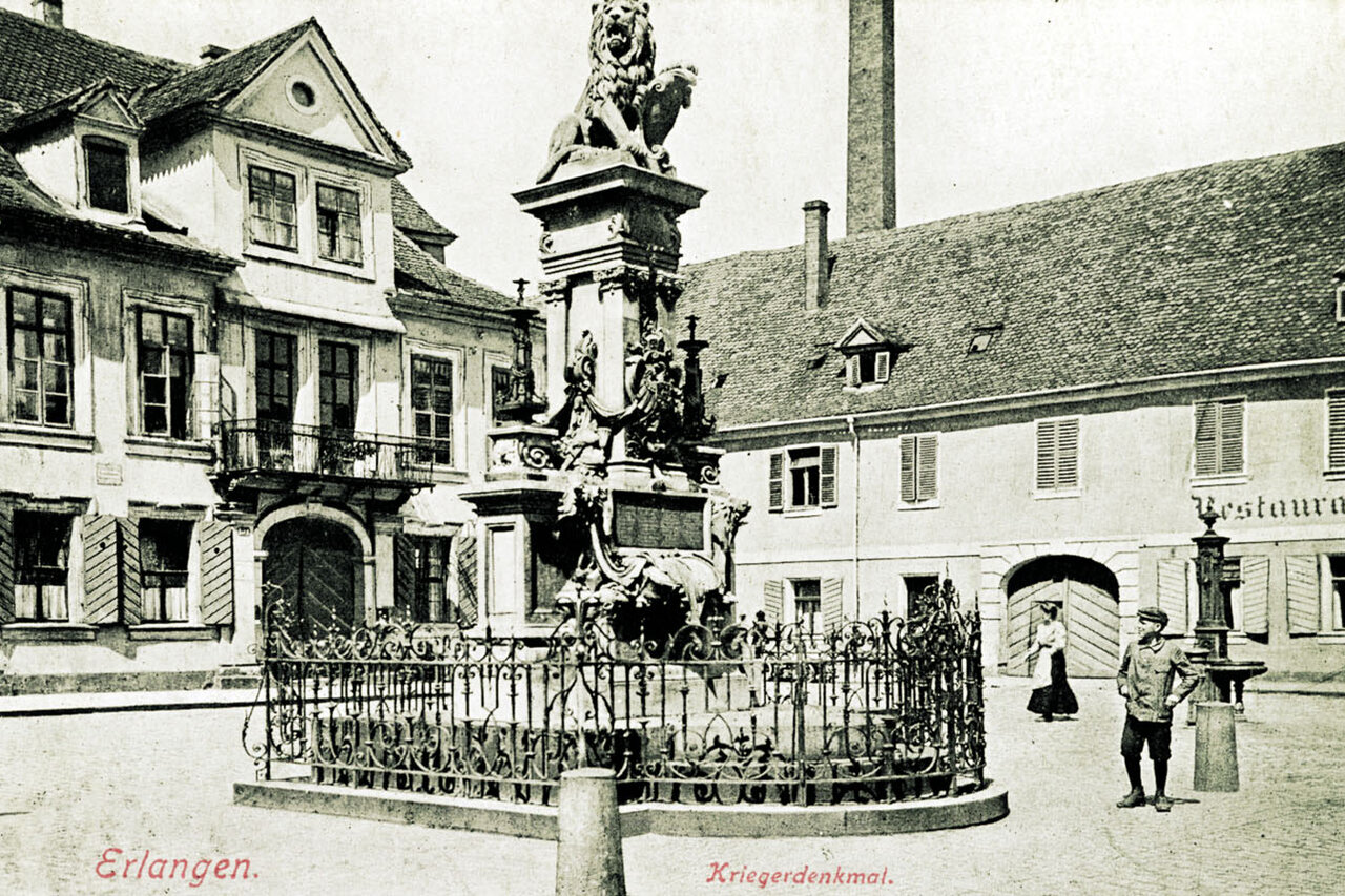 Historische Postkarte, im Zentrum das Kriegerdenkmal mit einem Löwen auf einem Platz, der von Häusern umgeben ist, unten die Aufschrift 'Erlagen Kriegerdenkmal'