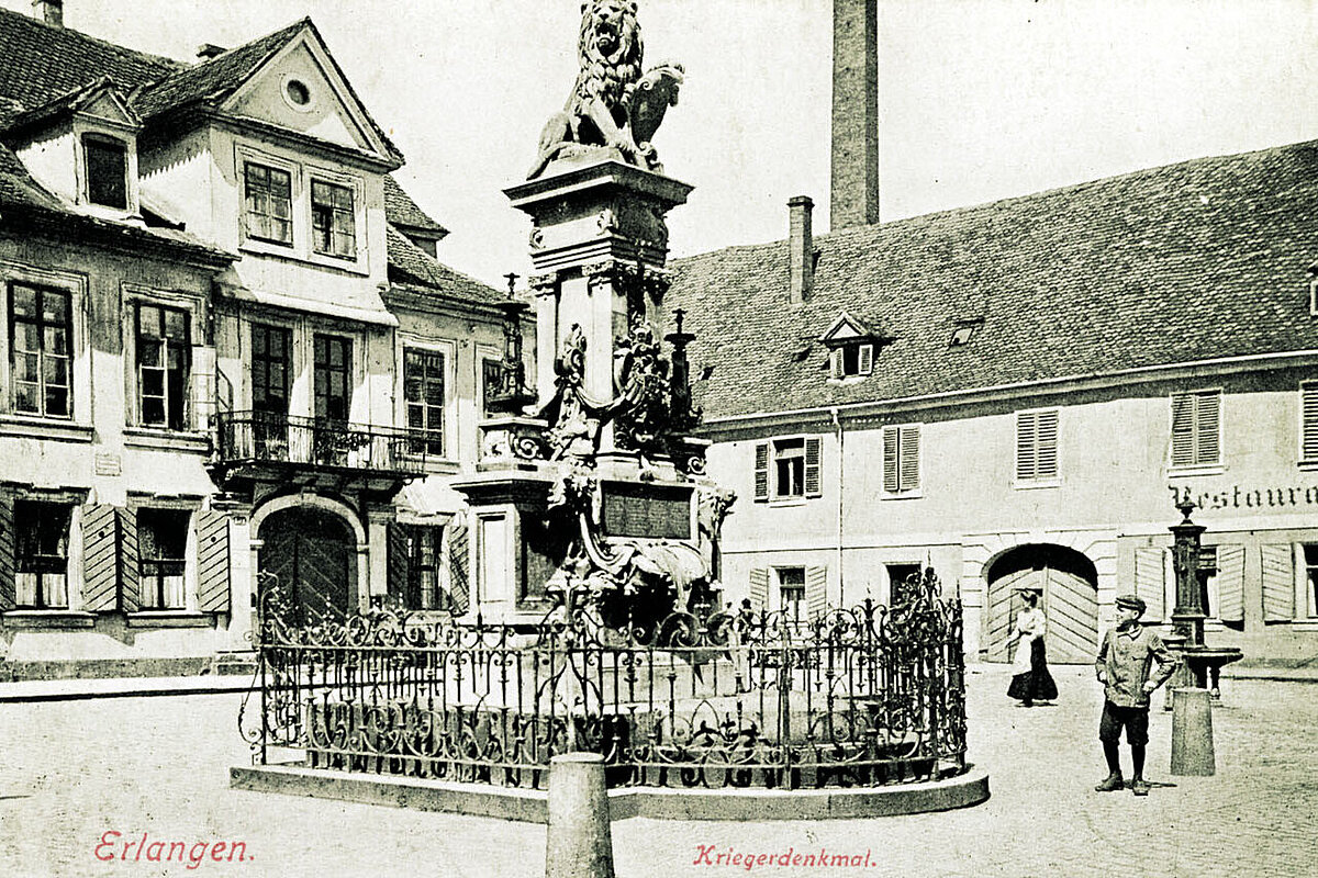 Historische Postkarte, im Zentrum das Kriegerdenkmal mit einem Löwen auf einem Platz, der von Häusern umgeben ist, unten die Aufschrift 'Erlagen Kriegerdenkmal'
