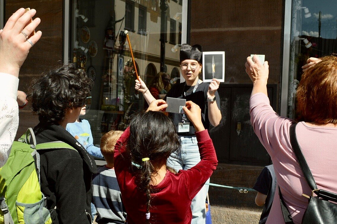 Rundgangsleiterin mit Hut und Zepter hält ein Bild hoch, vor ihr stehen Kinder und Erwachsene, die verschiedene Gegenstände in die Luft halten