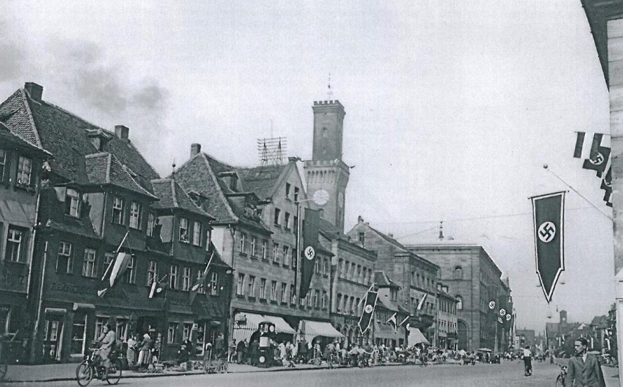 Schwarzweißaufnahme von einer großen Straße, über der Hakenkreuzfahnen hängen, im Hintergrund eine Häuserzeile und das Fürther Rathaus