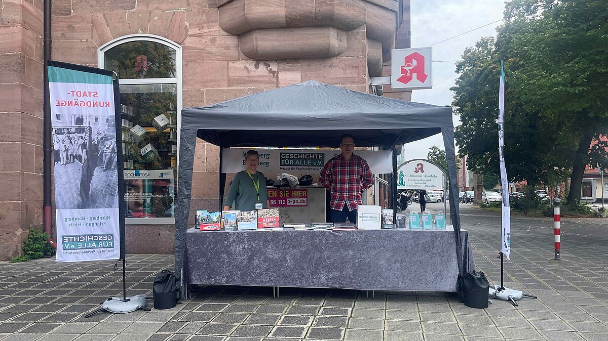 Ruth Papadopoulos und Markus Heidebroek am Infostand, GFA 2025. Infostand von Geschichte Für Alle mit einem Pavillon und zwei Beachflags vor einem einem Gebäude. Unter dem Pavillon stehen zwei Personen hinter dem Tisch, auf dem Tisch sind Bücher und Flyer.