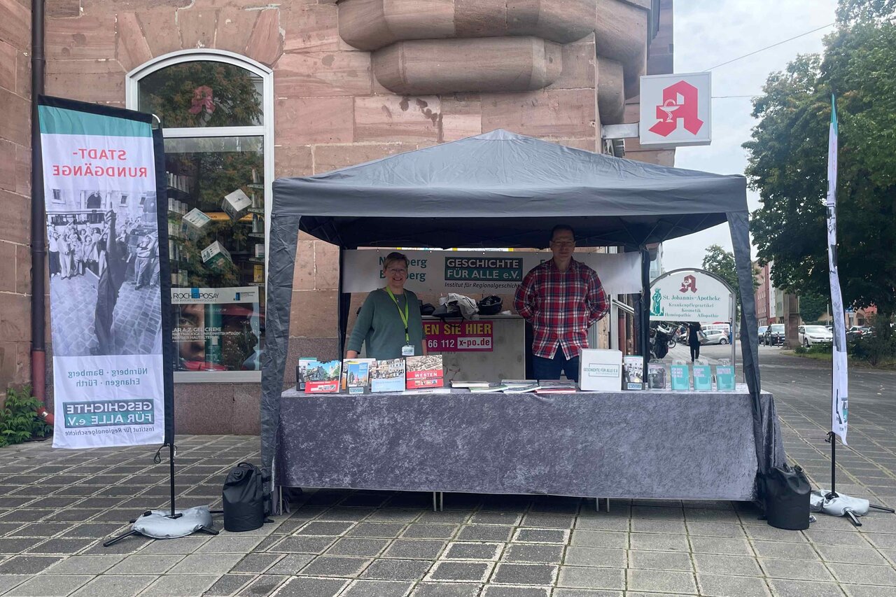 Infostand von Geschichte Für Alle mit einem Pavillon und zwei Beachflags vor einem einem Gebäude. Unter dem Pavillon stehen zwei Personen hinter dem Tisch, auf dem Tisch sind Bücher und Flyer.