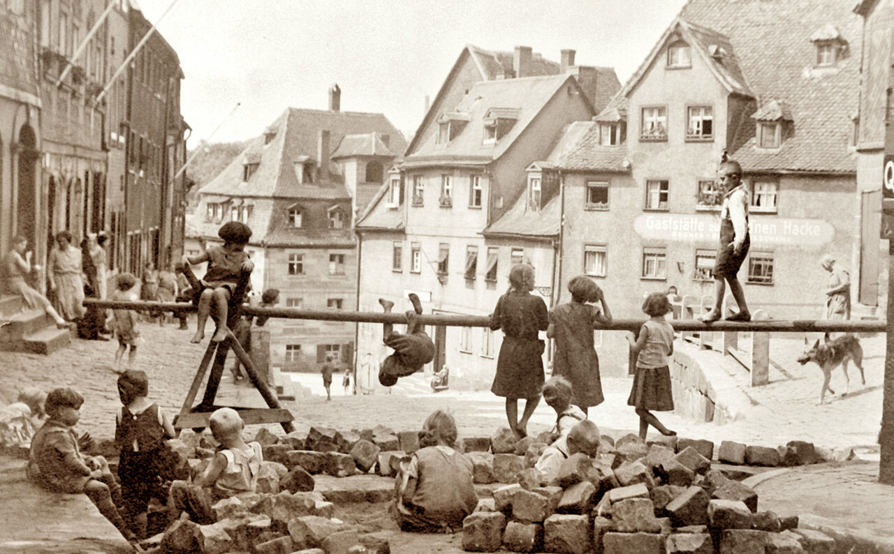 Schwarzweißaufnahme von spielenden Kindern in einer Straße, im Hintergrund Häuserzeilen