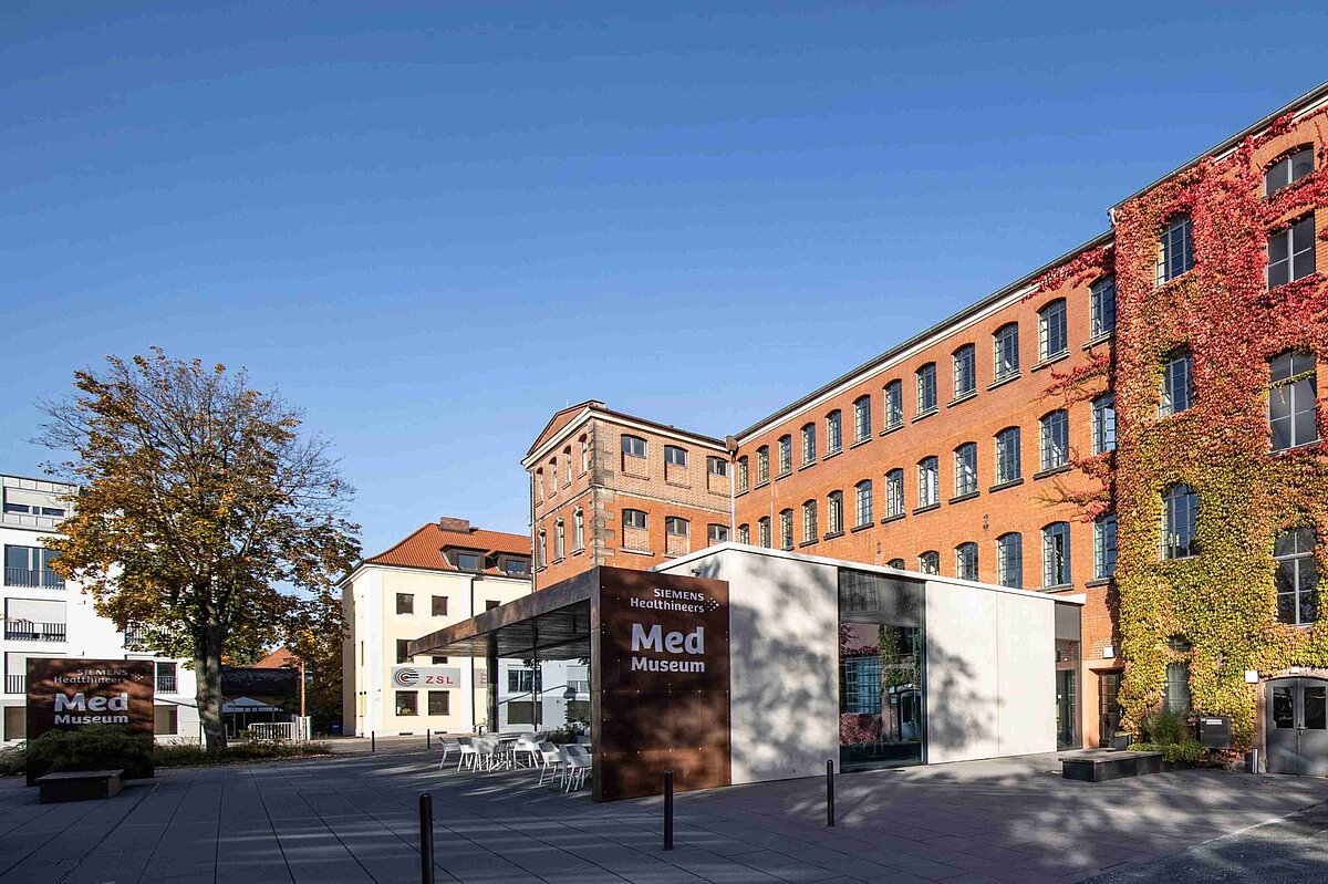 Außenseite des Siemens Healtheneers MedMuseums. In der Mitte der kleine Anbau mit dem Eingangsbereich, auf dem groß MedMuseum steht und vor dem einige Stühle stehen, dahinter der große Backsteinbau des Museums.