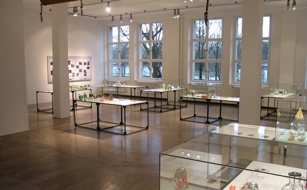 Ausstellungsraum des Museums Bayerische Metallwarenfabrik mit großen Fenstern, Holzboden und Glasvitrinen mit Exponaten