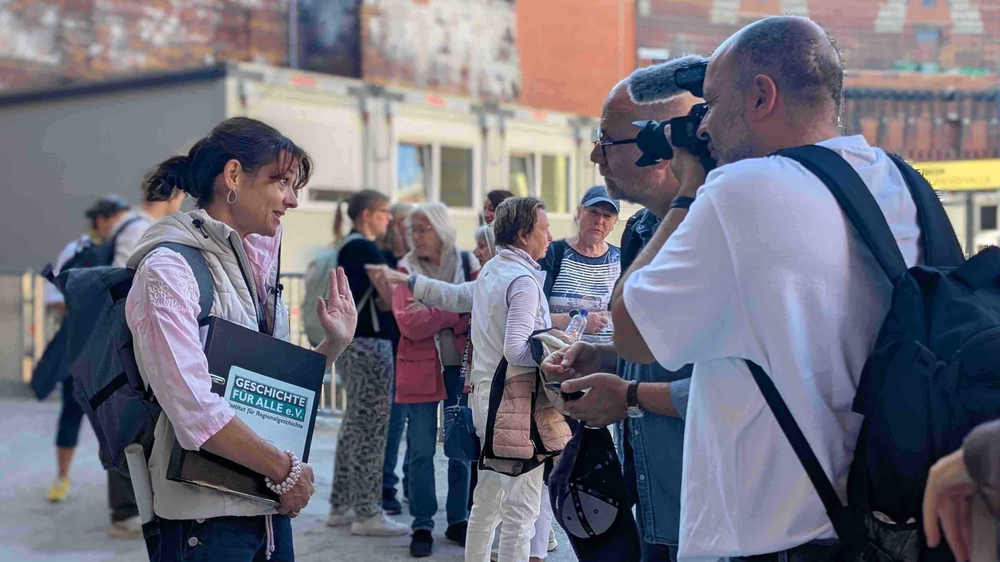 Person mit Ordner unter dem Arm unterhält sich mit einer Person, sie werden von einem Kameramann gefilmt. Im Hintergrund weitere Menschen vor einem Ziegelsteingebäude.  