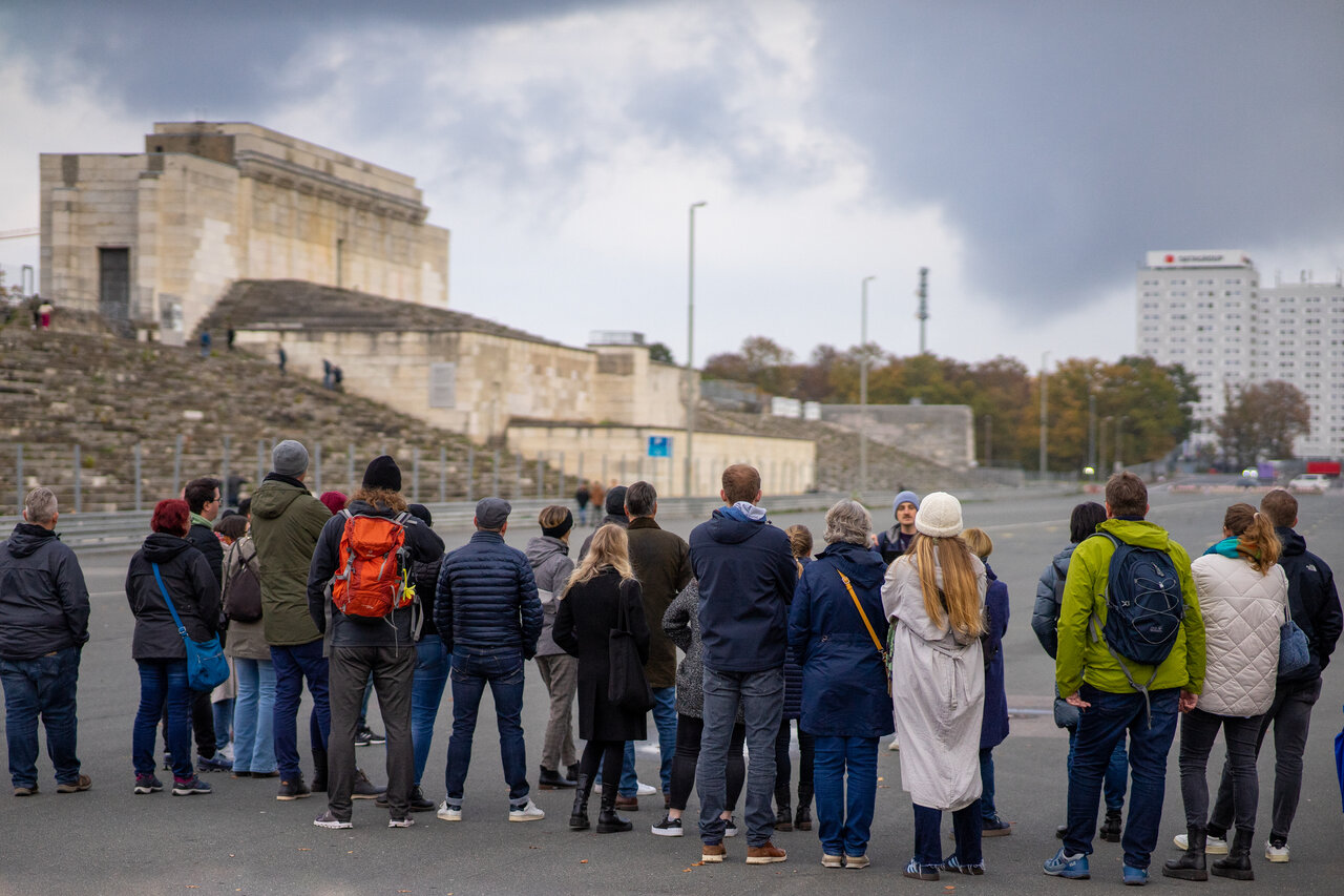 Eine Gruppe Menschen von hinten Fotografiert, im Hintergrund unscharf die Zeppelintribüne auf dem ehemaligen Reichsparteitagsgelände