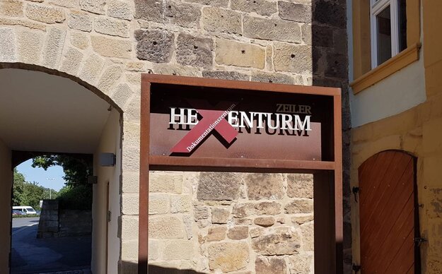 Schild mit der Aufschrift 'Hexenturm' vor einer Sandsteinmauer, links ein Torbogen