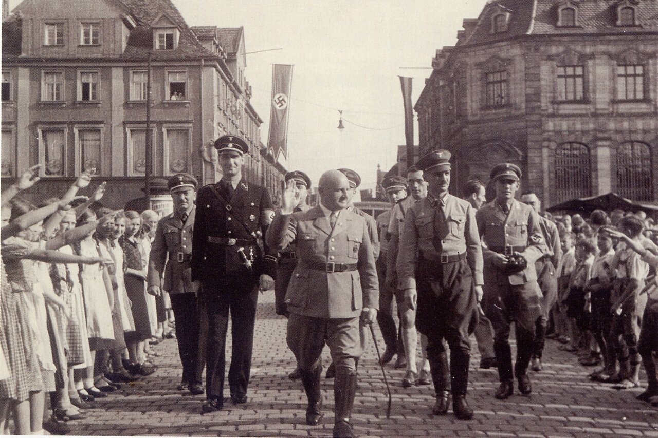 Schwarzweißaufnahme von hochrangigen Nationalsozialisten in Uniform, umgeben von Kindern, die den Hitlergruß machen, im Hintergrund Hakenkreuzfahnen zwischen den Barocken Häusern Erlangens