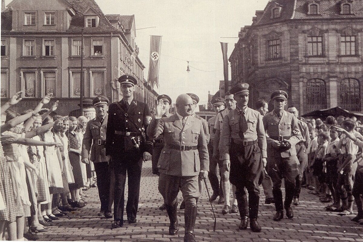 Schwarzweißaufnahme von hochrangigen Nationalsozialisten in Uniform, umgeben von Kindern, die den Hitlergruß machen, im Hintergrund Hakenkreuzfahnen zwischen den Barocken Häusern Erlangens