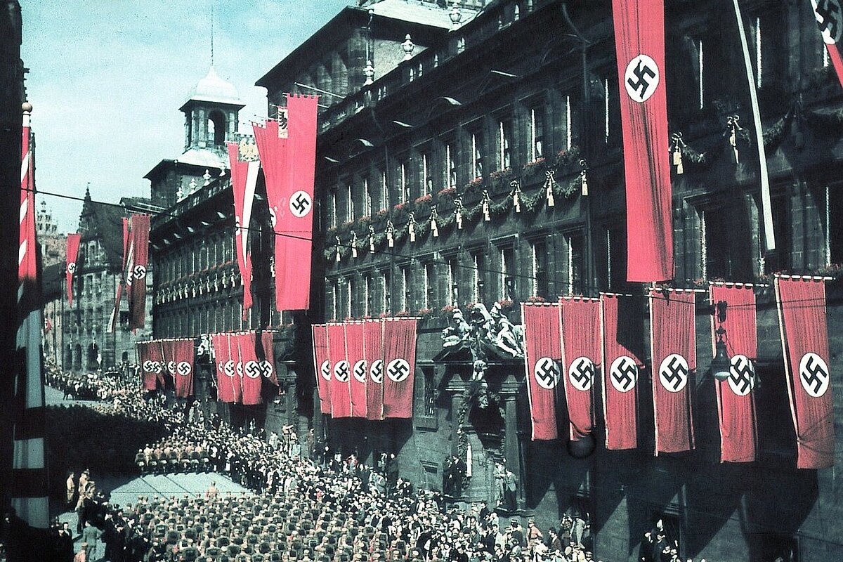 Kolorierte Fotograf des Nürnberger Rathauses, an dessen Fassade rote Hakenkreuzfahnen hängen, in der Straße davor Aufmarsch von Nationalsozialisten