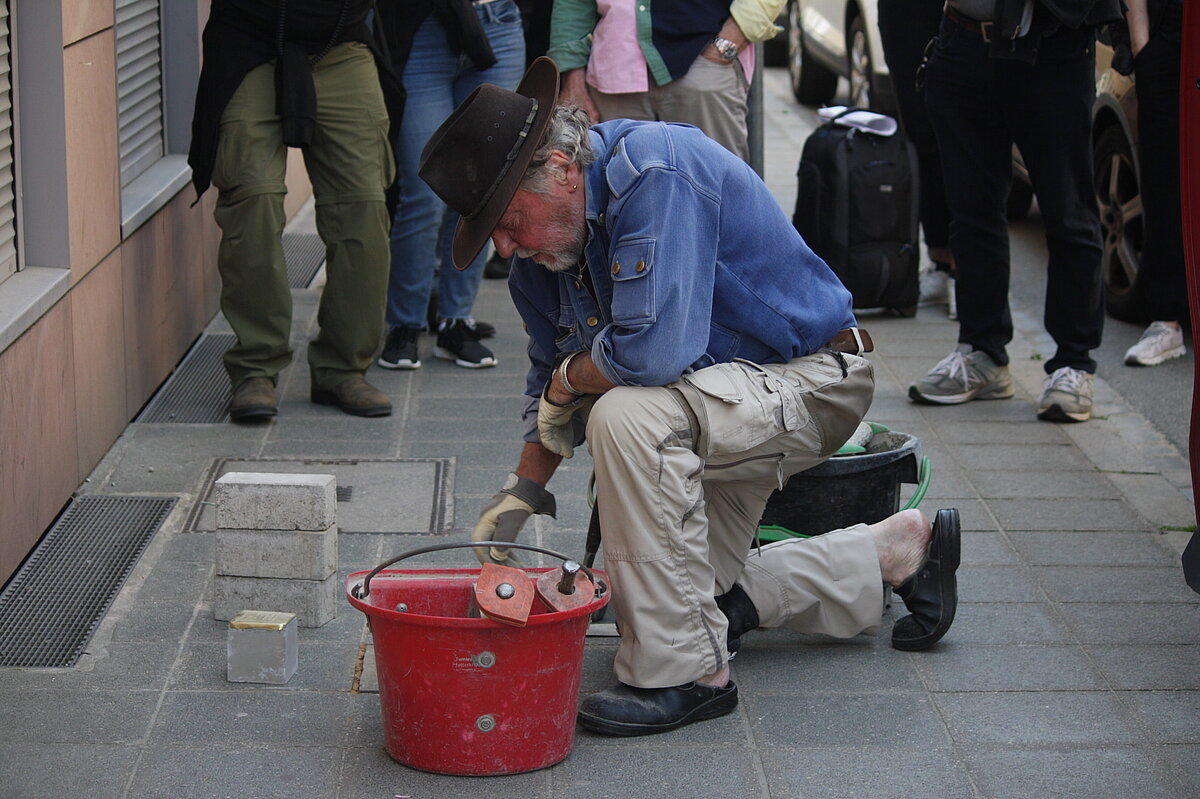 Ein Mann mit blauem Hemd und dunklem Hut kniet auf dem Gehsteig, neben sich einen roten Eimer, in der Hand einen Spachtel, im Hintergrund eine kleine Menschenmenge.