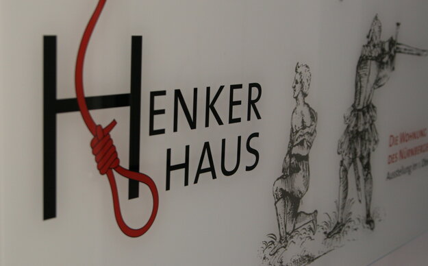 Eingangstafel des Museums Henkerhau, im Fokus ein großes 'H' mit einer stilisierten Galgenschlinge, daneben unscharf die historische Darstellung einer Enthauptung durch das Schwert