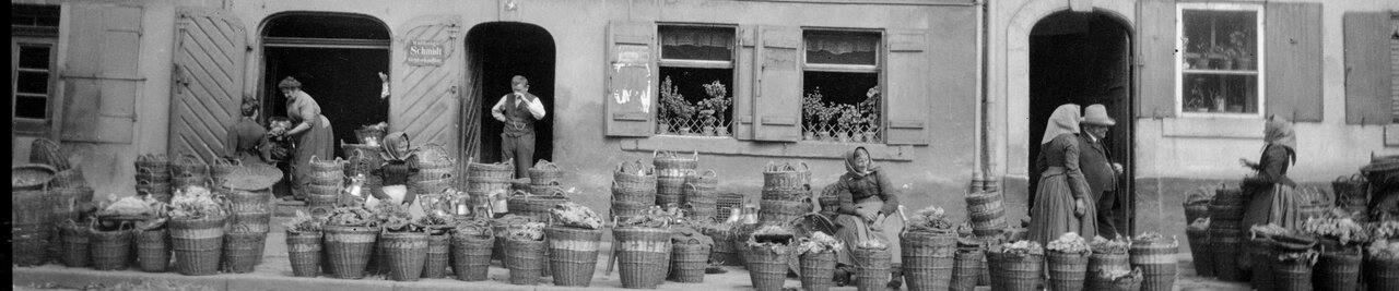 Marktszene mit vielen Körben am Straßenrand sowie Verkäuferinnen und Passante vor einer Häuserzeile, Schwarzweißfotografie von 1910