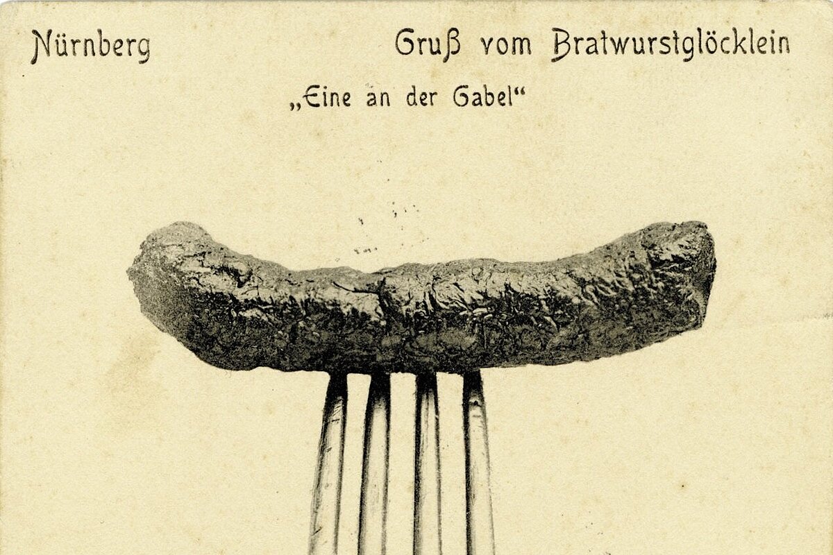 Historische Postkarte mit einer auf einer Gabel aufgespießten Bratwurst vor neutralem Hintergrund und der Aufschrift 'Nürnberg Gruß vom Bratwurstglöcklein Eine an der Gabel'