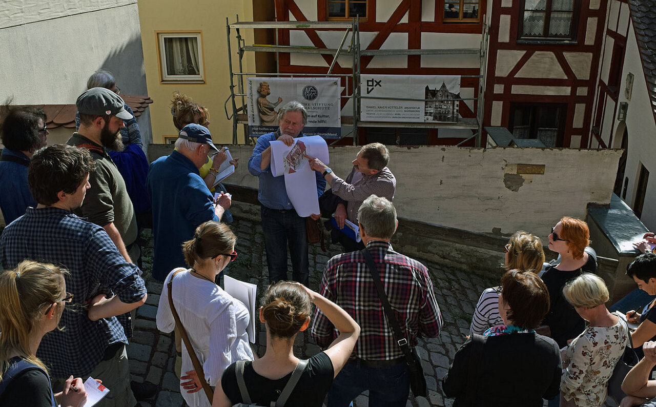 Eine Gruppe Menschen steht um zwei Männer, der eine hält einen Bauplan, der andere deutet darauf, im Hintergrund ein Fachwerkhaus mit Gerüst