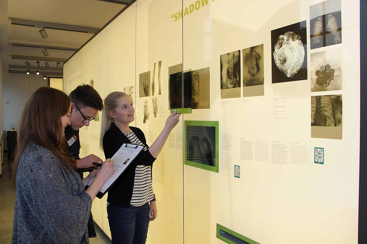 Drei Jugendliche, die sich der Wand ihnen gegenüber zuwenden, an der Röntgenaufnahmen verschiedener Körperteile hängen. Ein Mädchen hält ein Bild daneben, um sie zu vergleichen, die anderen machen sich Notizen.