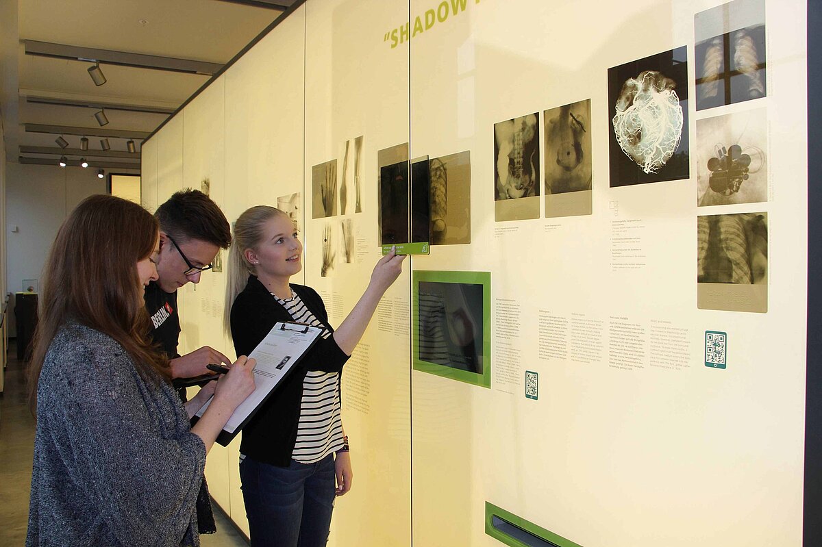 Drei Jugendliche, die sich der Wand ihnen gegenüber zuwenden, an der Röntgenaufnahmen verschiedener Körperteile hängen. Ein Mädchen hält ein Bild daneben, um sie zu vergleichen, die anderen machen sich Notizen.