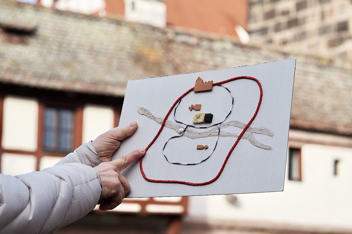Ein Tastmodell der Nürnberger Altstadt wird von einer Hand senkrecht nach oben gehalten, während es die andere Hand abtastet, im Hintergrund ein Fachwerkhaus
