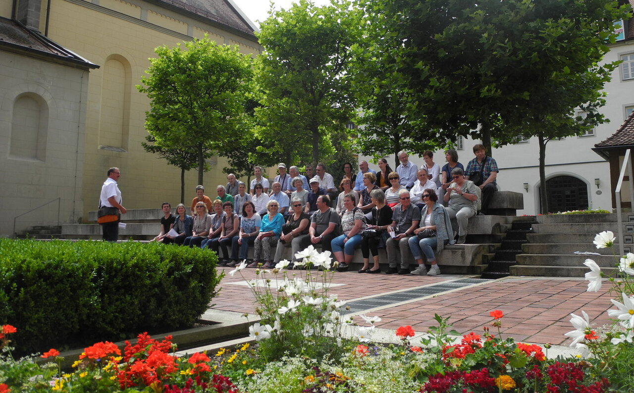Im Vordergrund ein Blumenbeet im Sonnenschein, im Hintergrund eine Gruppe Menschen auf einer Treppe sitzend