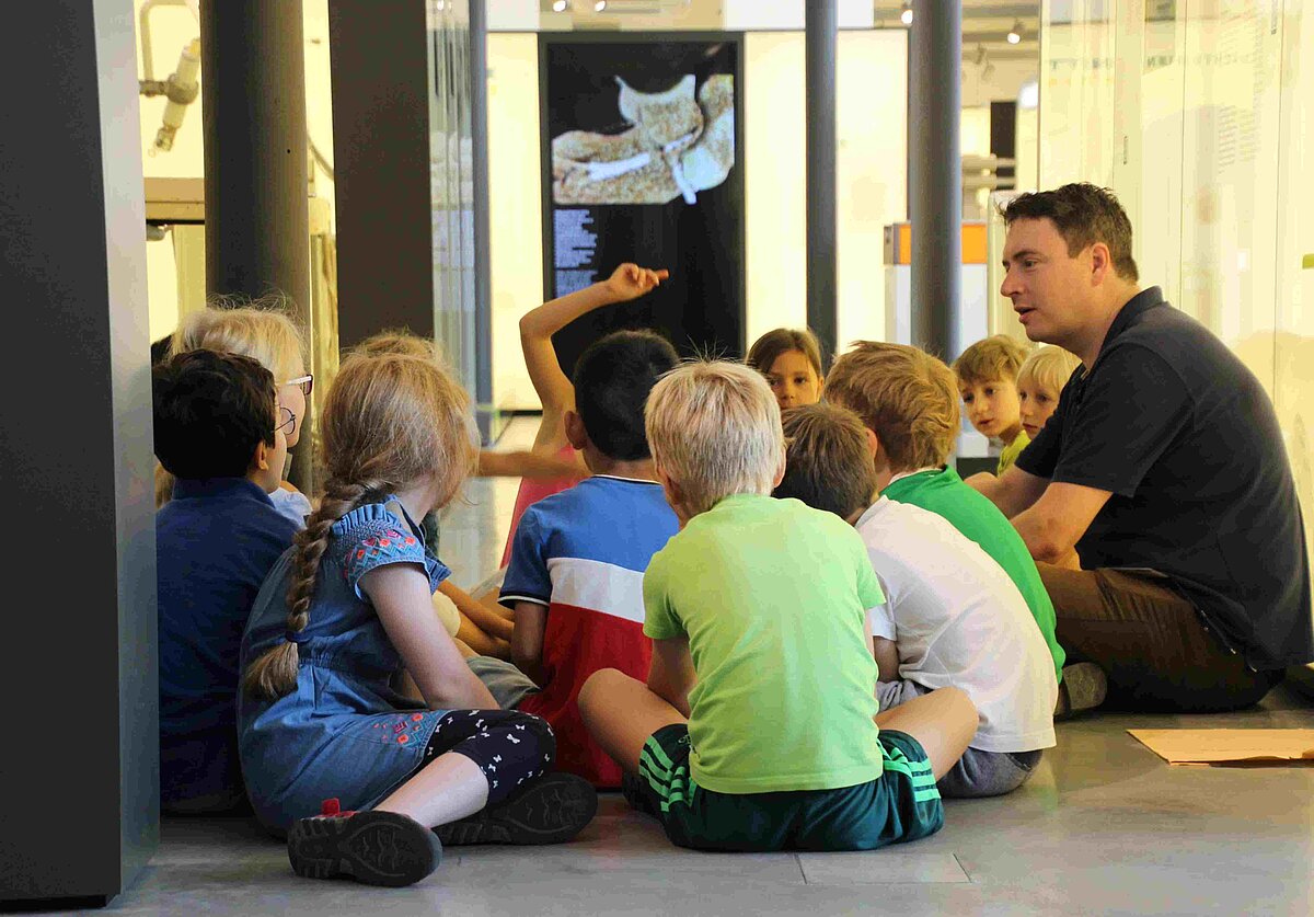 Eine Gruppe Kinder im Grundschulalter und ein erwachsener Mann sitzen im Kreis. Die meisten Kinder haben dem Betrachter den Rücken zugewandt und ihren Blick auf den Mann gerichtet. Ein Kind meldet sich.