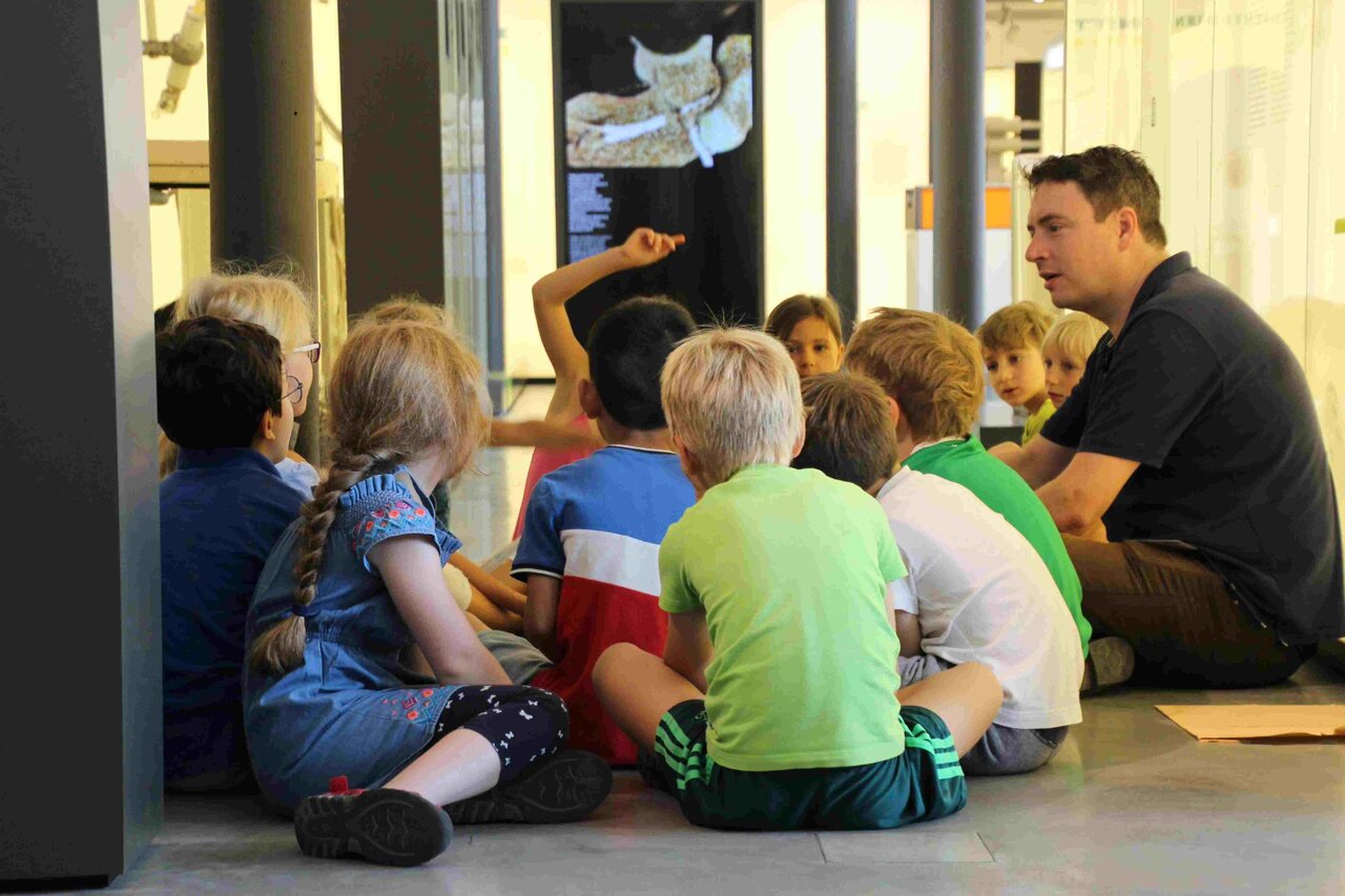 Eine Gruppe Kinder im Grundschulalter und ein erwachsener Mann sitzen im Kreis. Die meisten Kinder haben dem Betrachter den Rücken zugewandt und ihren Blick auf den Mann gerichtet. Ein Kind meldet sich.
