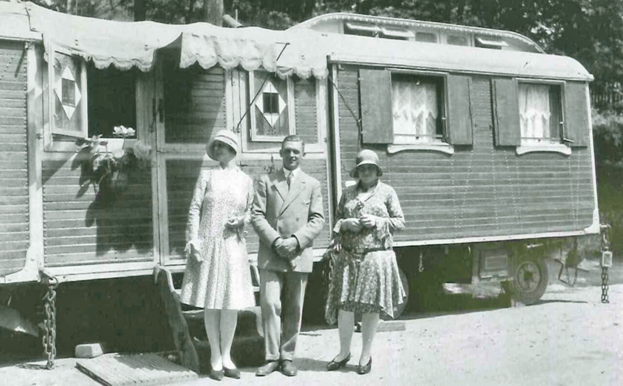 Historische Schwarzweißfotografie eines Wohnwagens aus Holz mit geöffneten Fenstern, davor ein Mann und zwei Frauen in vornehmer Kleidung