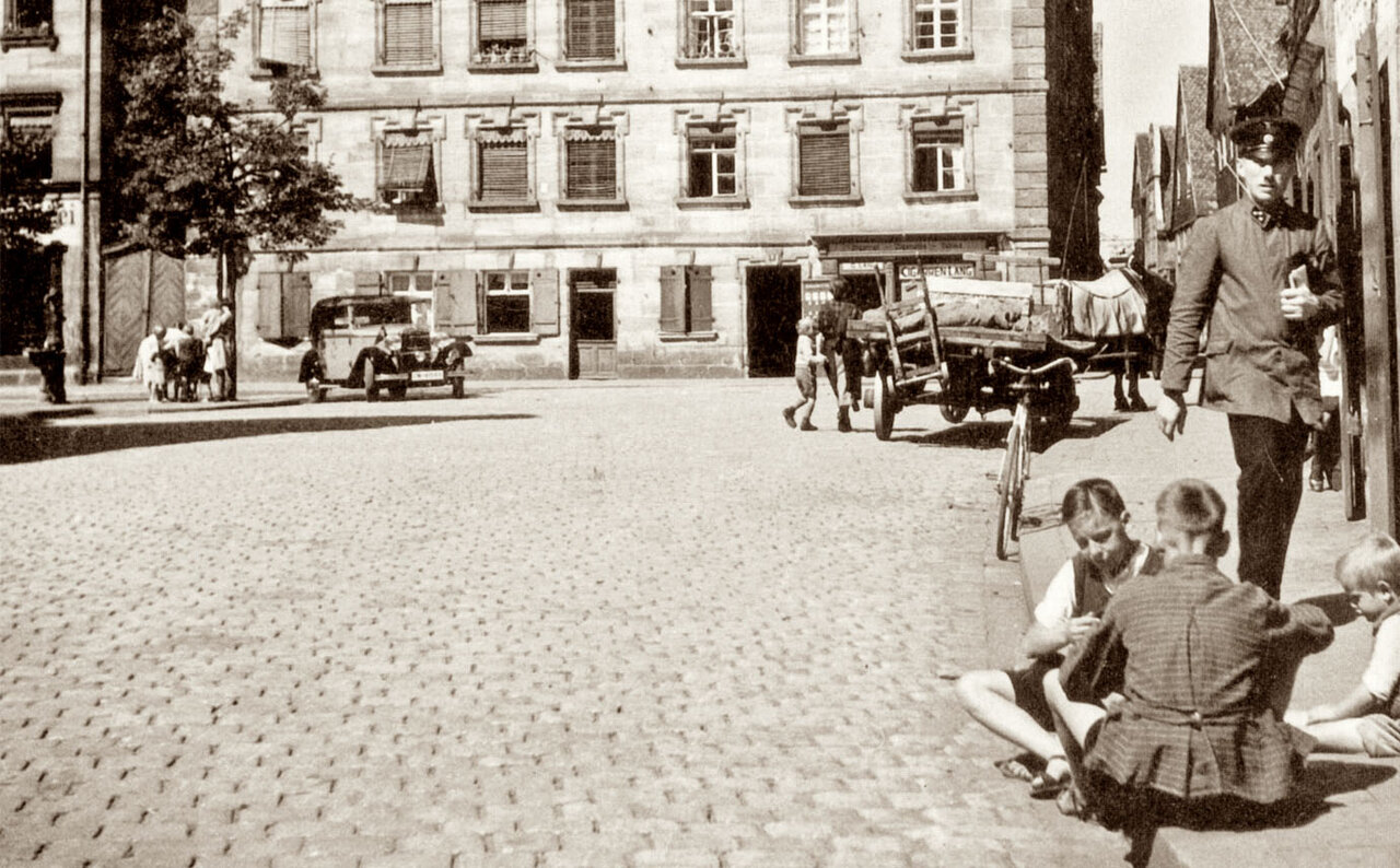 Schwarzweißaufnahme von einer gepflasterten Straße, links ein Automobil, rechts auf dem Boden sitzende Kinder und ein stehender Mann, im Hintergrund ein großes Haus