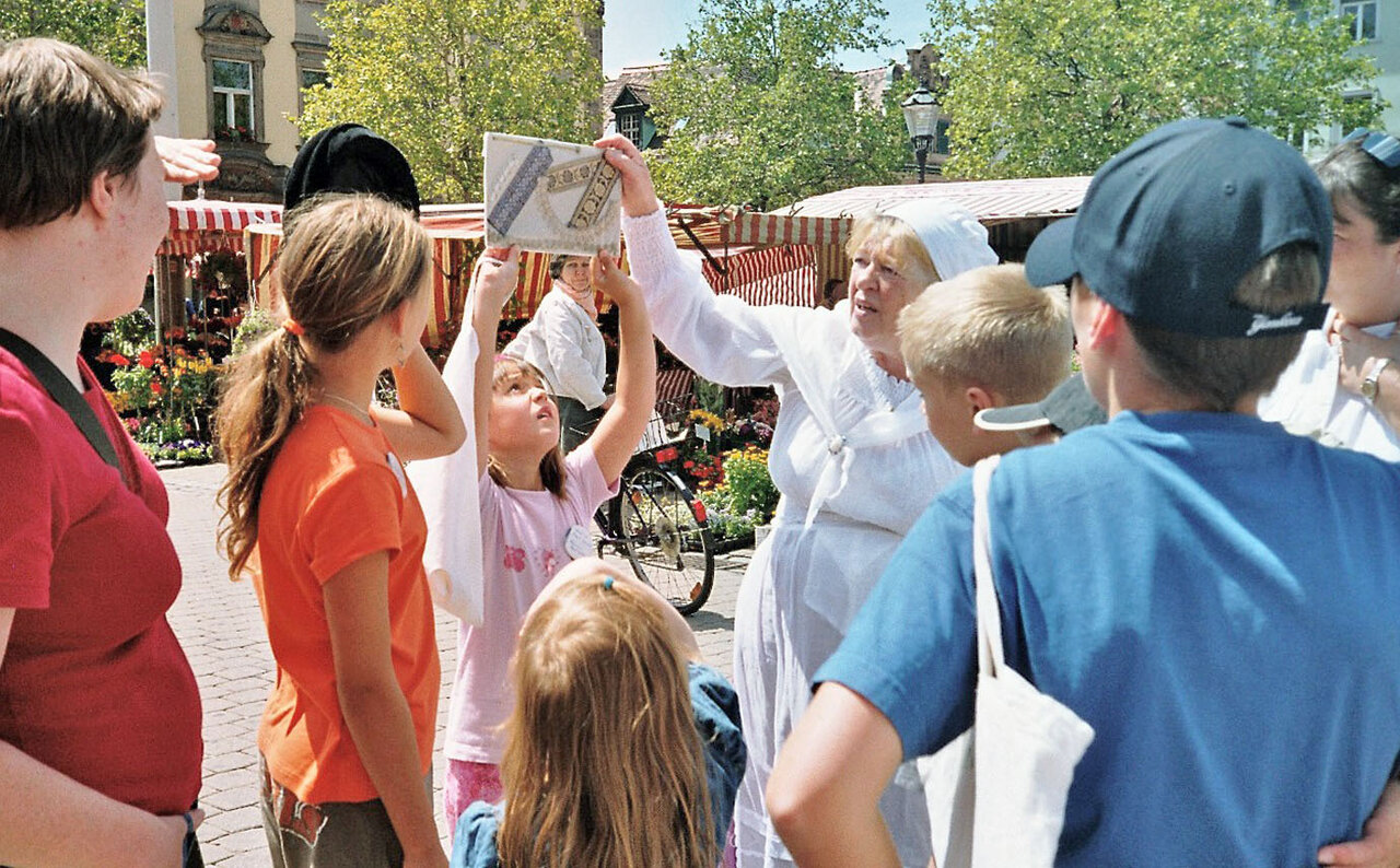 Gruppe von Kindern schaut auf ein Bild, das ein Mädchen hochhält, neben ihr die Rundgangsleiterin im historischen Kostüm einer Magd, im Hintergrund der Marktplatz