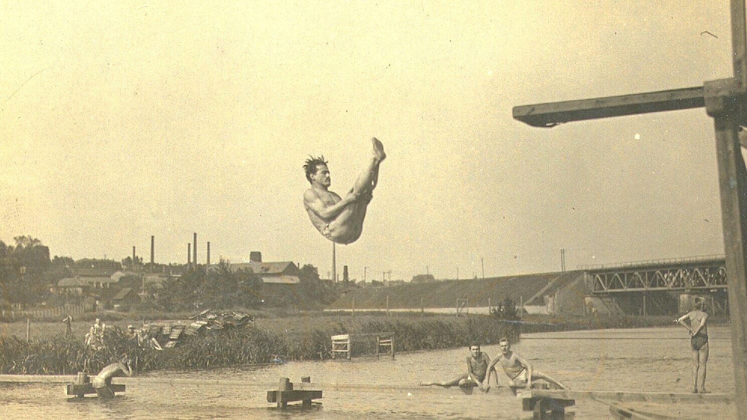 Historische Fotografie von einem Mann, der im Freibad Pulversee einen Rückwärtssalto von einem Sprungturm macht, im Hintergrund andere Badegäste, um 1920