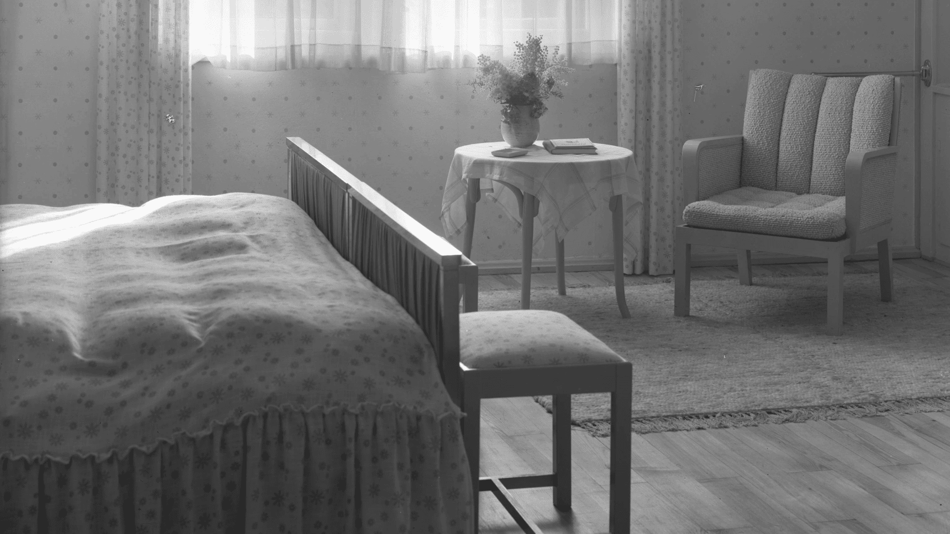 Schwarzweißaufnahme eines schlichten Gästezimmers mit Bett, Sessel, Tisch und Blumenstrauß, um 1940