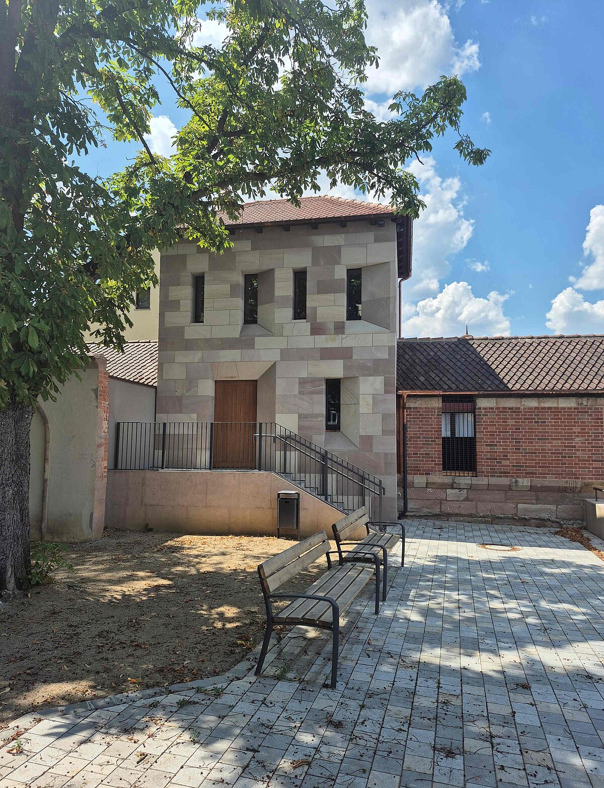 Fotografie Turm Blaues G im Hintergrund mit einem Teil des Wehrgangs. Im Vordergrund auf der rechten Seite ein gepflasterter Boden, eine Sitzbank. In den linken Bildrand ragen die belaubten Zweige eines Baumes herein.