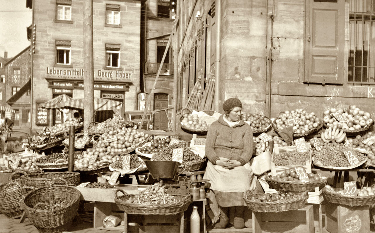 Historische Fotografie von einer Marktfrau, die zwischen gefüllten Körben sitzt, im Hintergrund Steinhäuser