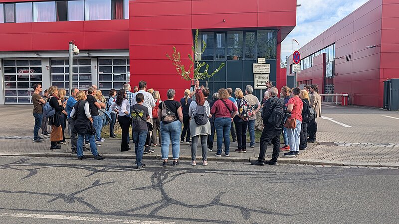 Eine Menschengruppe steht um den Rundgangsleiter und lauscht vor einem roten Gebäude dem kostenlosen Jubiläumsrundgang
