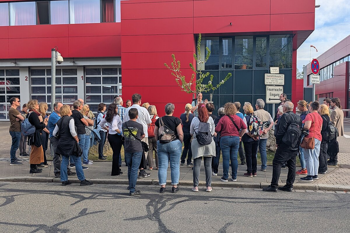 Eine Menschengruppe steht um den Rundgangsleiter und lauscht vor einem roten Gebäude dem kostenlosen Jubiläumsrundgang