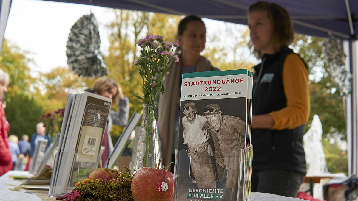 Christine Maß und Jennifer Oevermann auf dem Apfelmarkt © Ron Hübner, 2022. Flyer des Vereins 'Geschichte Für Alle' neben einem Blumenstrauß und herbstlicher Tischdekoration, im Hintergrund unscharf zwei Mitarbeiterinnen des Vereins