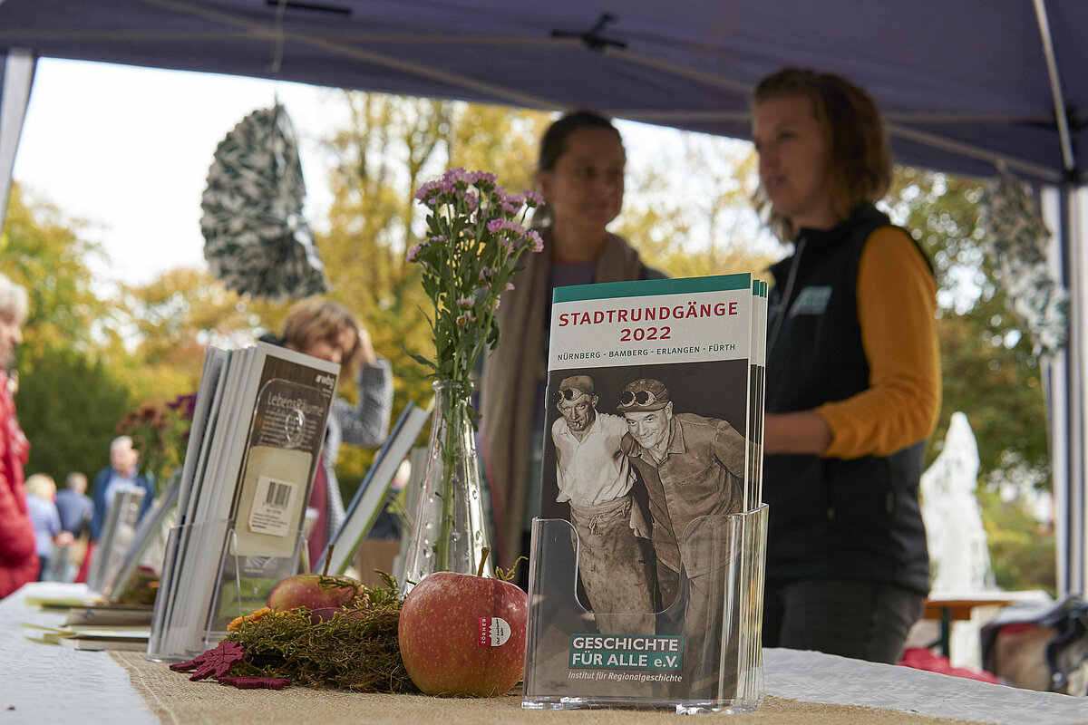 Christine Maß und Jennifer Oevermann auf dem Apfelmarkt © Ron Hübner, 2022. Flyer des Vereins 'Geschichte Für Alle' neben einem Blumenstrauß und herbstlicher Tischdekoration, im Hintergrund unscharf zwei Mitarbeiterinnen des Vereins