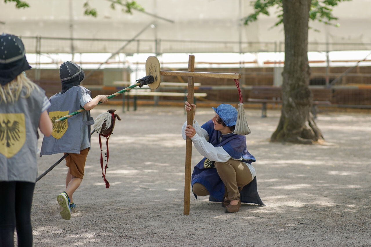 Rudngangsleiterin und Kinder in mittelalterlichen Kostüme spielen ein Ritterturnier nach