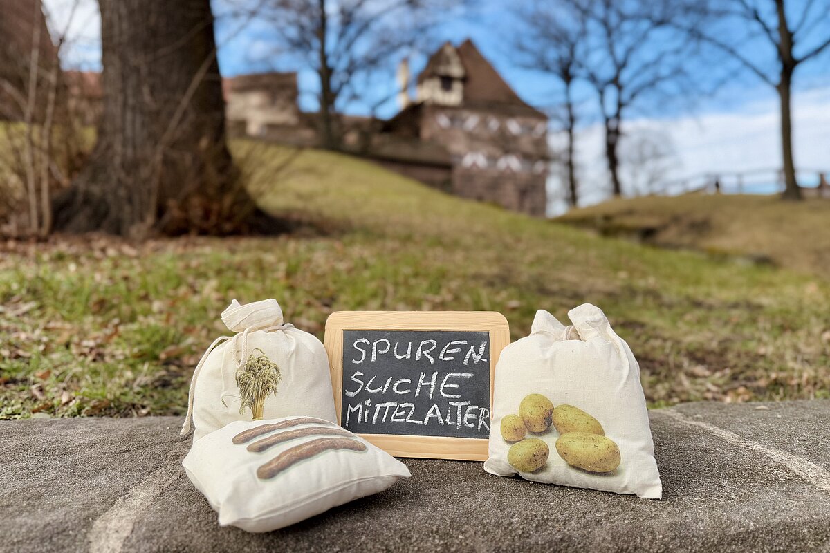 Auf einem Sandsteinblock liegen drei kleine Leinensäckchen und eine kleine Schreibtafel mit der Aufschrift 'Spurensuche Mittelalter', im Hintergrund eine Wiese mit Bäumen sowie ein Fachwerkhaus