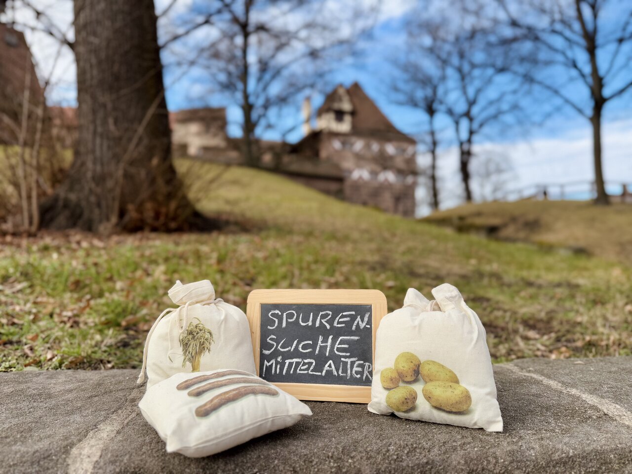 Auf einem Sandsteinblock liegen drei kleine Leinensäckchen und eine kleine Schreibtafel mit der Aufschrift 'Spurensuche Mittelalter', im Hintergrund eine Wiese mit Bäumen sowie ein Fachwerkhaus