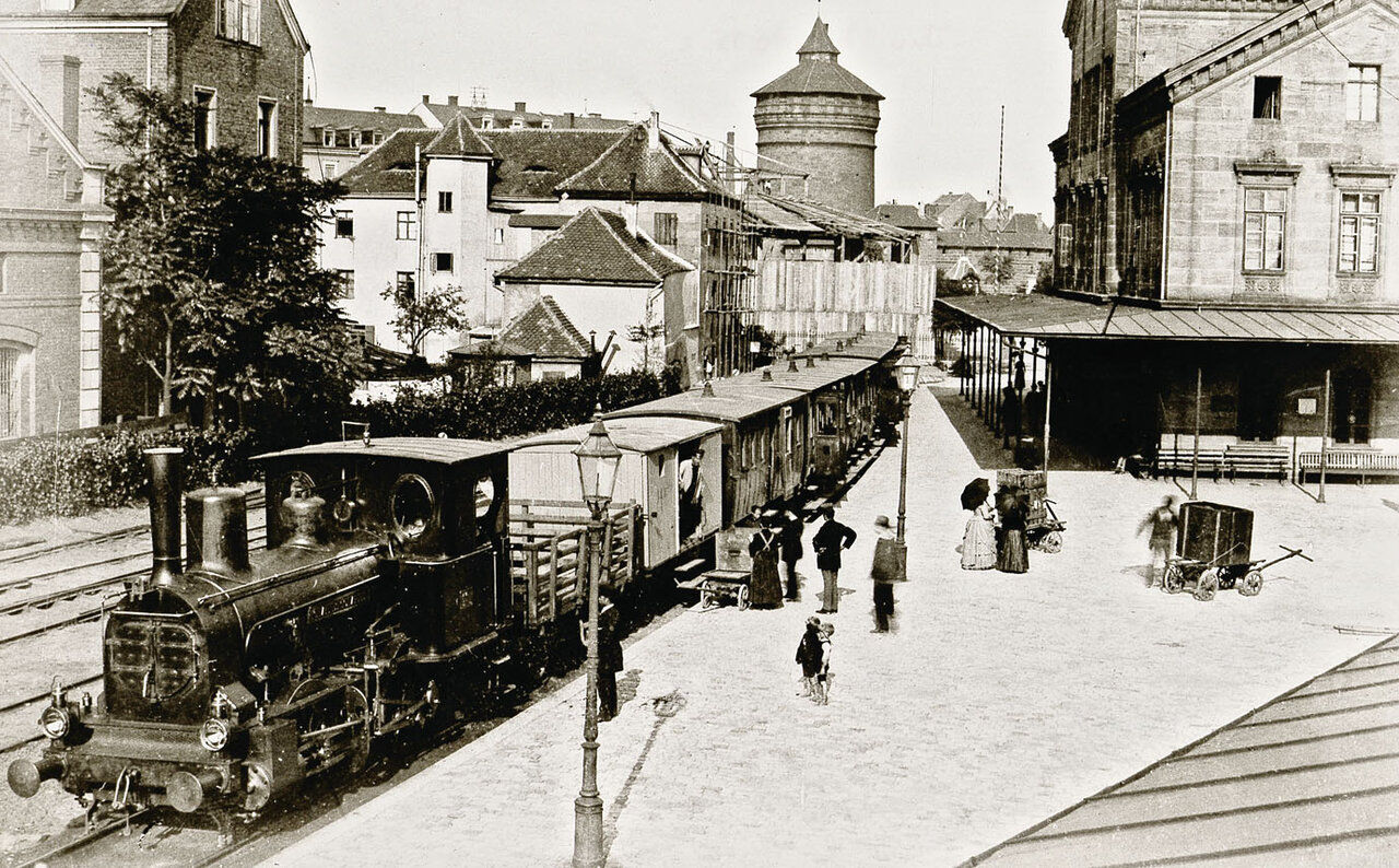 Schwarzweißweißaufnahme von einer Dampflok mit Waggons in der Fürther Straße, im Hintergrund Häuser und Türme