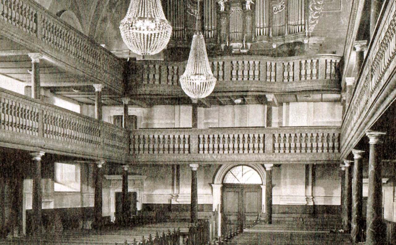 Schwarzweißaufnahme vom Innenraum einer Kirche mit Holzbänken, zwei Emporen übereinander, Kristallkronleuchtern an der Decke und einer großen Orgel