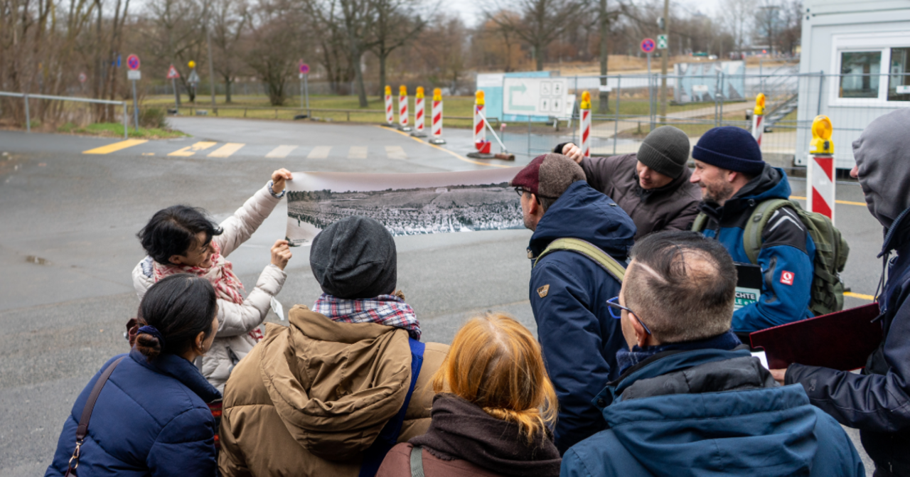 Eine Gruppe Auszubildender ist von hinten zu sehen, sie schauen auf ein ausgerolltes Bild einer Schwarzweißaufnahme, im Hintergrund unscharf eine Baustelle