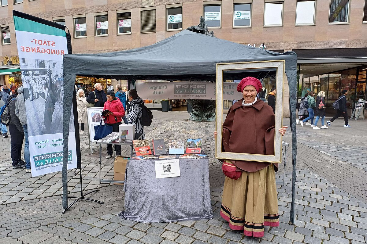 Vor dem Infostand aus einem Tisch mit Büchern und Infomaterial des Vereins unter einem grauen Pavillon steht eine Frau in mittelalterlichem Kostüm und schaut durch einen hölzernen Rahmen.
