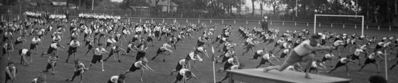 Eine Gruppe von Schülern macht auf einem Sportplatz eine Übung, am rechten Bildrand ein Vorturner auf einem Podest, Schwarzweißaufnahme um 1930