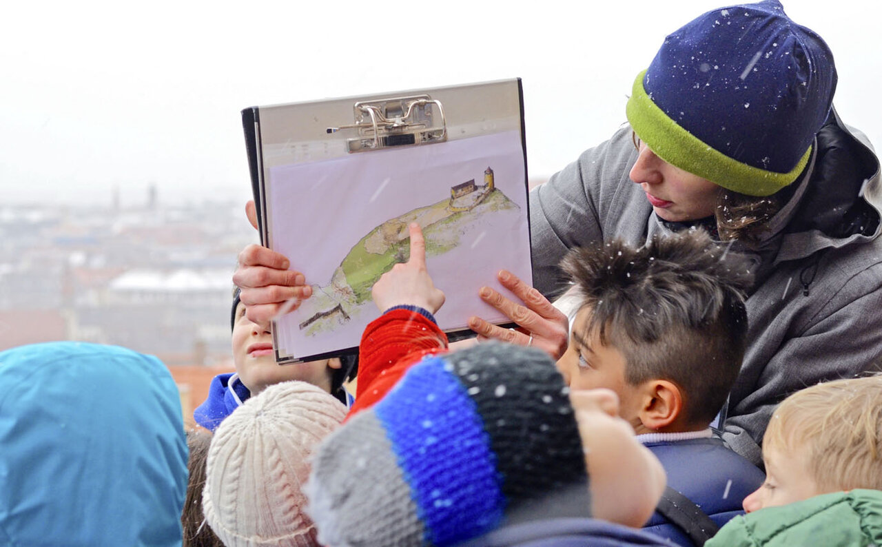 Rundgangsleiterin hält ein Bild der Burg hoch, die Kinder sind von hinten zu sehen, hören zu und eines zeigt auf das Bild